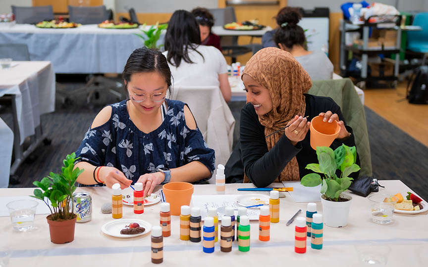 Two students are seated at a table chatting as they use an array of paint colors to paint plant pots.