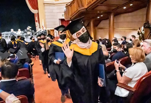 graduates in academic regalia smile and wave as they walk up a theatre aisle