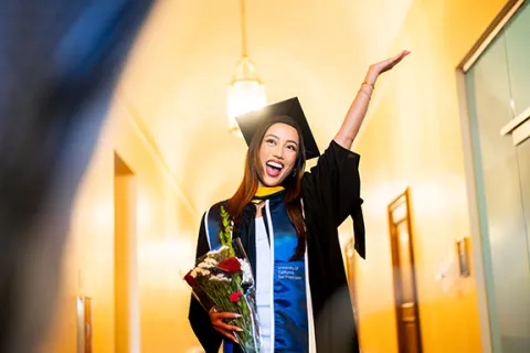 a grad in regalia holding a bouquet of flowers raises her arm in celebration