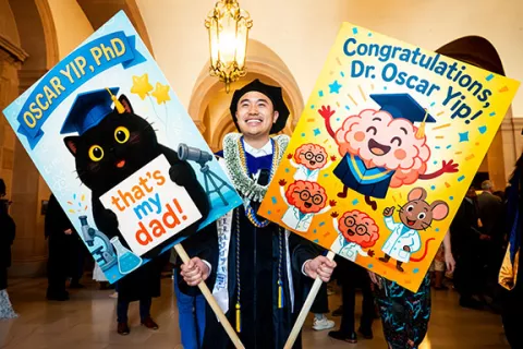 a grad in regalia holds two large celebratory signs
