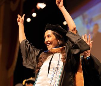 a graduate in academic regalia holds her arms up in celebration after being hooded at commencement