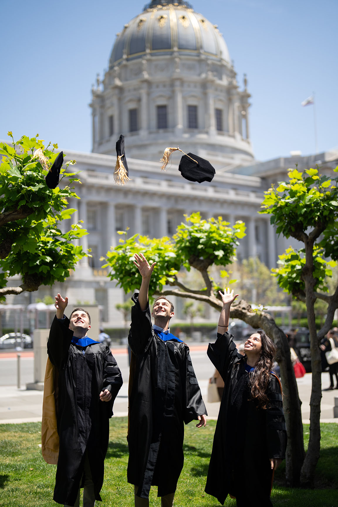three new grads toss their caps in the air with SF City Hall in the background