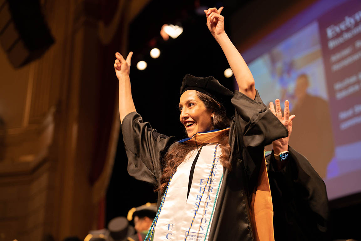a graduate raises her hands in celebration after being hooded on stage