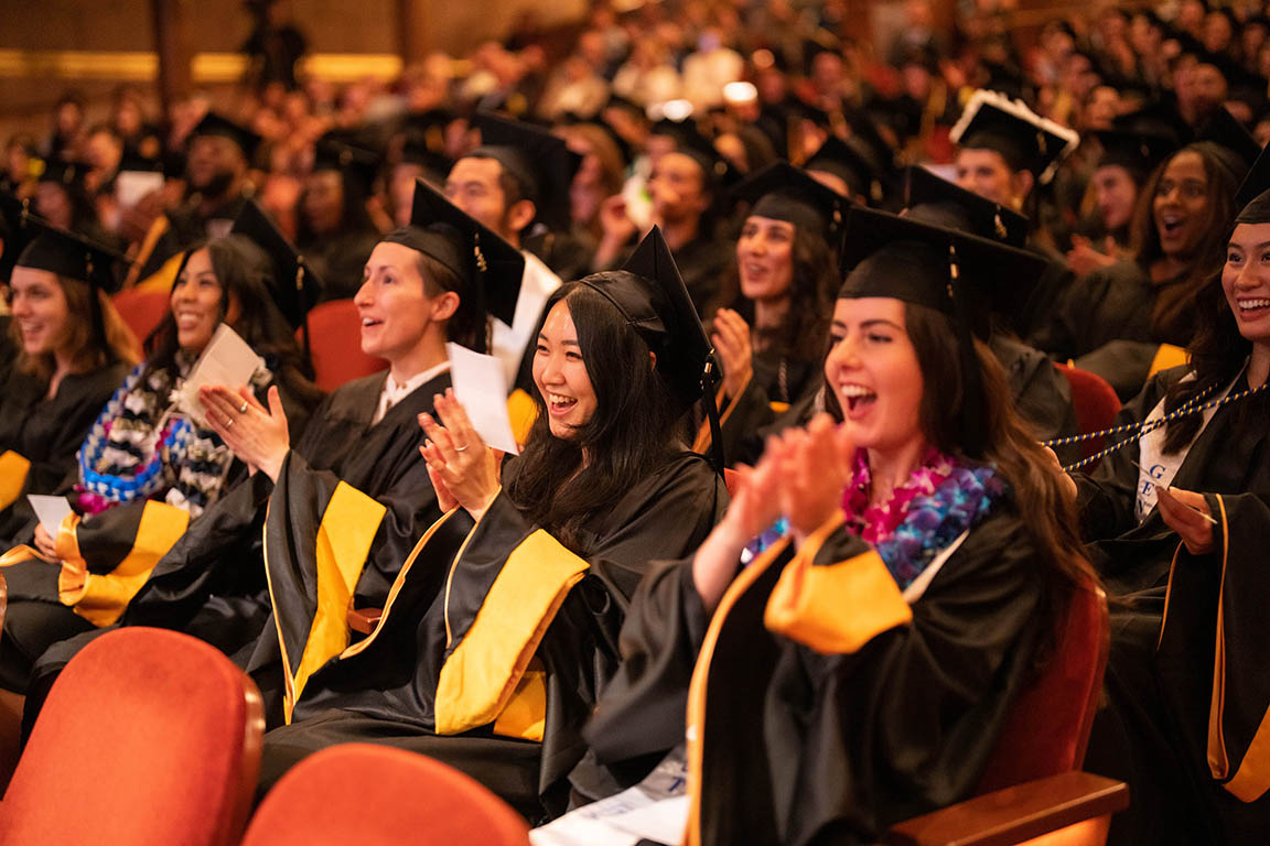 Class of 2023 graduates, in full regalia, clap and smile in the audience at the Graduate Division Commencement Ceremony
