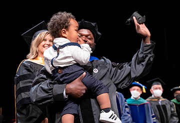 Antoine Johnson, PhD, hold his young son in his arms and waves to the audience after being hooded by Professor Aimee Medeiros, who looks on
