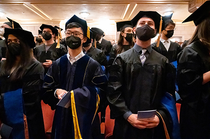 PhD grads in academic regalia stand in the audience awaiting the start of the ceremony after the academic procession