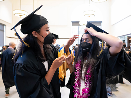 two graduates in commencement regalia prepare for procession; one wears a magenta flower lei