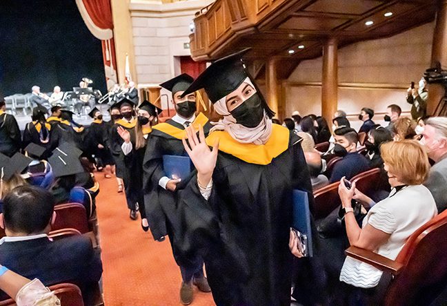graduates in academic regalia smile and wave as they walk up a theatre aisle
