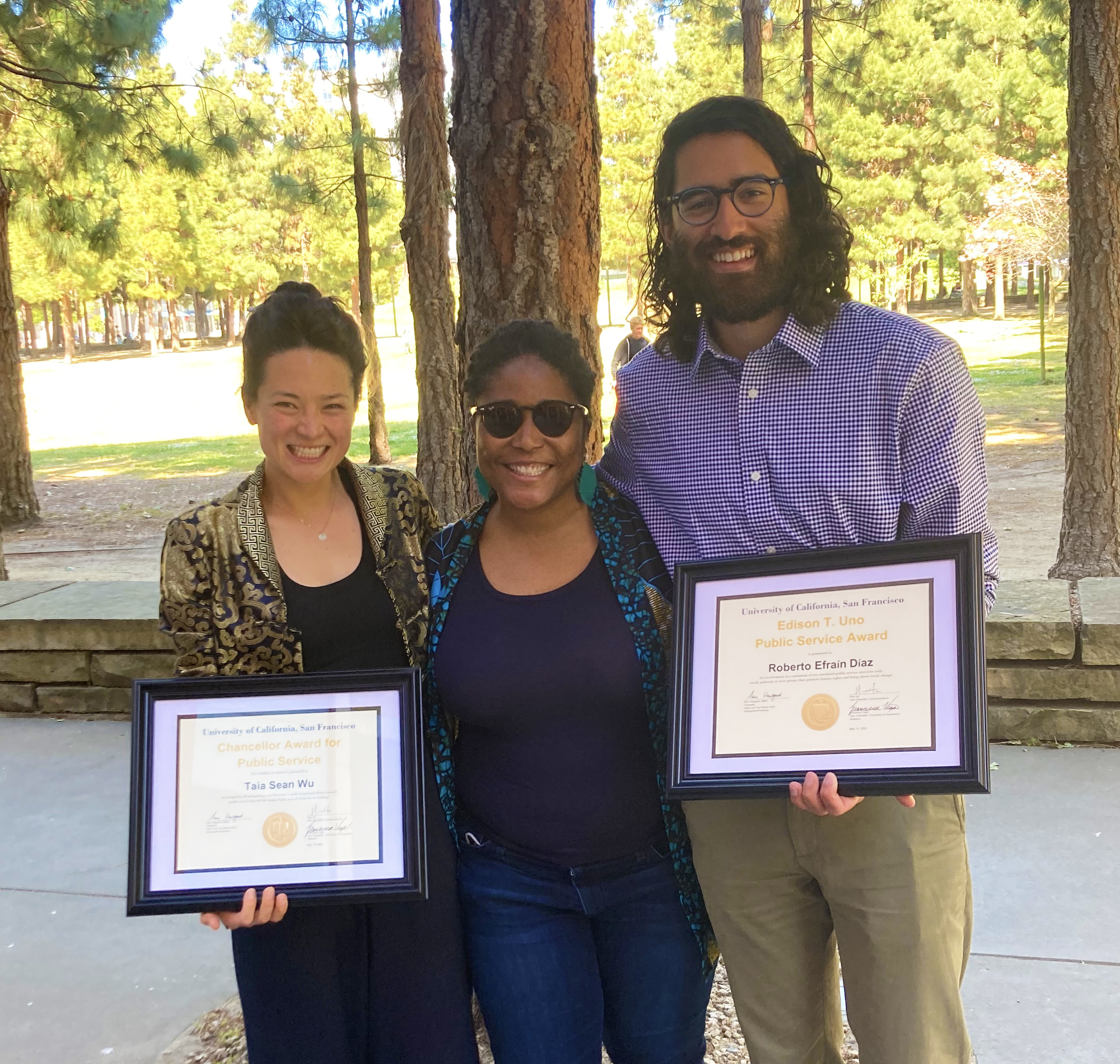 Taia Wu, D'Anne Duncan, and Robbie Diaz smile for a photo under pine tree. Taia and Robbie hold their framed awards